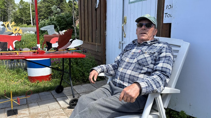 Alain Landry, assis sur une chaise en plastique devant son atelier, à côté d'une table présentant des girouettes et créations en métal.