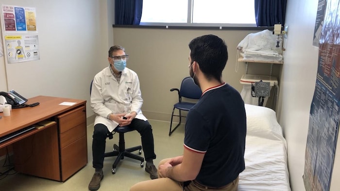 Un médecine portant un masque parle à un patient assis sur un lit.