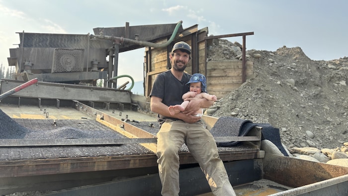 Un homme et son bébé sont assis sur une immense machine servant à extraire l'or des sédiments.