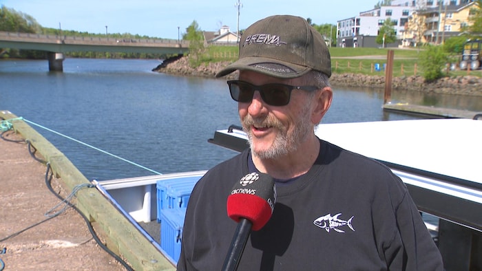 Un homme portant des lunettes fumées et une casquette est interviewé sur un quai, devant un bateau de pêche.