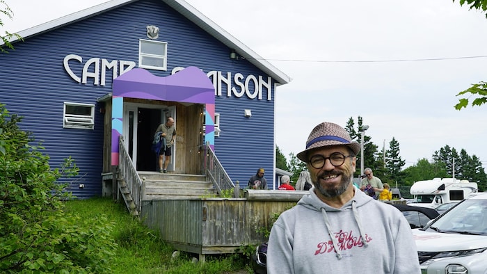 Le directeur du Festival en chanson porte un chapeau  et sourit à l'objectif devant un bâtiment du camp en chanson.