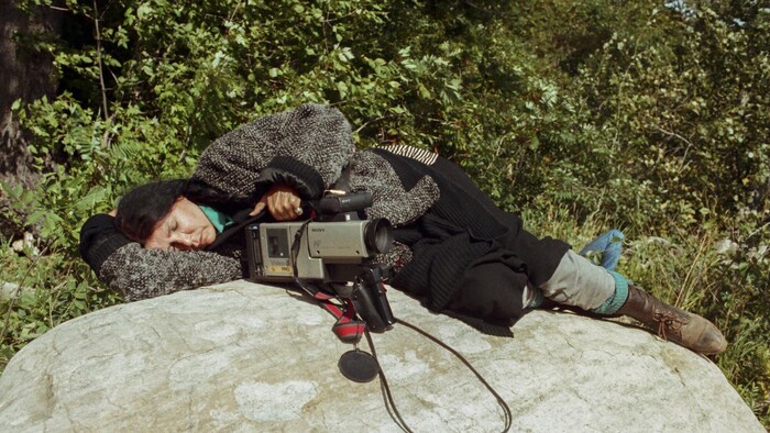 Alanis Obomsawin est étendue les yeux fermés sur une roche, un caméscope à la main.