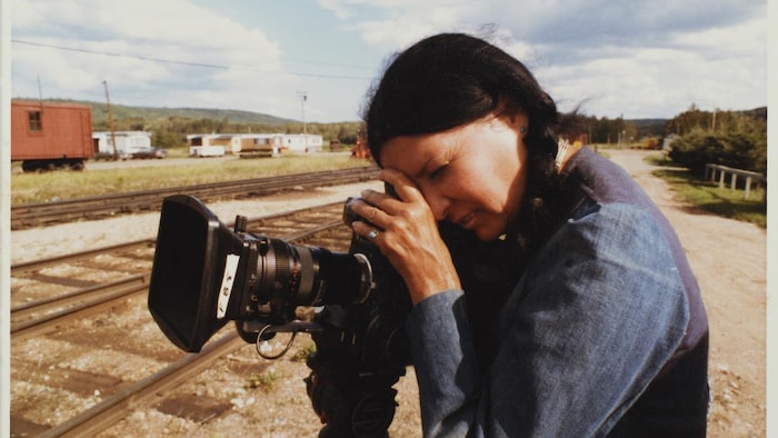 Devant un chemin de fer, Alanis Obomsawin regarde dans un caméscope.