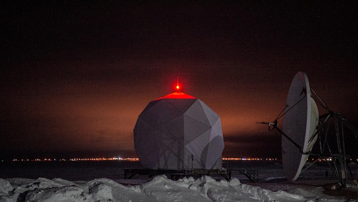 Un grande sphère avec une lumière allumée sur le dessus, à côté d'un radar dans un décor hivernal la nuit. 