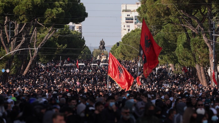 Une foule de personnes sont réunies sur une place près d'une statue, certains brandissent des drapeaux albanais.
