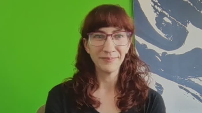 Le sourire aux lèvres, Janetta McKenzie porte des lunettes à monture rouge et a de longs cheveux auburn. Elle se trouve devant un fond vert uni avec un tableau abstrait en arrière-plan. Elle regarde la caméra.