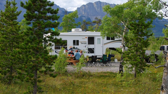 Quatre campeurs sont installés à une table de pique-nique, devant une caravane installée dans un terrain de camping de montagne.