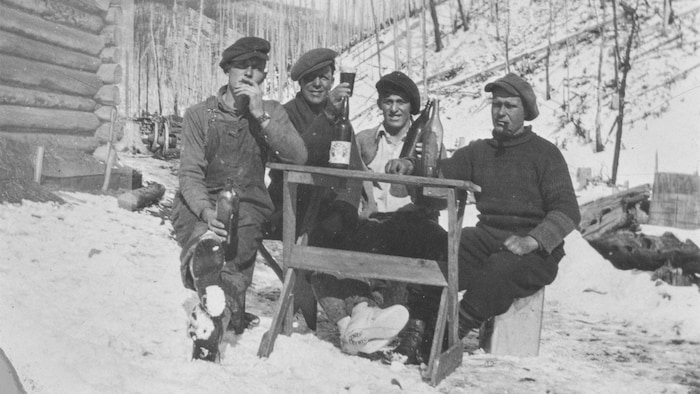 Quatre hommes assis dehors, à côté d'une cabane en bois rond.