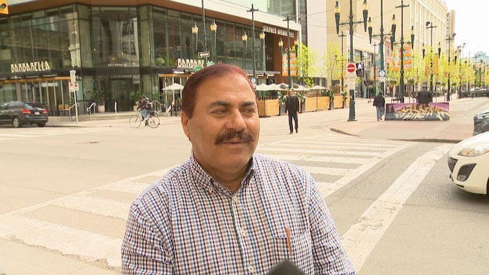 Un homme souriant aux cheveux courts bruns et arborant une moustache se tient au premier plan lors d'une entrevue à l'extérieur.