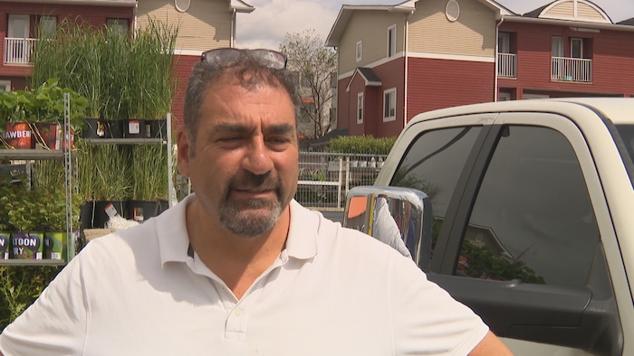 Un homme se tient à l'extérieur, entouré à gauche par des étagères remplies de plantes en pot et de grands plants d'herbe, et à droite par une camionnette blanche.
