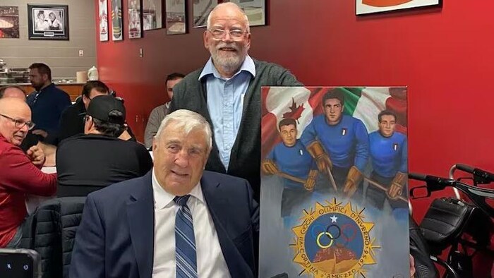 Aldo Maniacco, assis, alors qu'il recevait un tableau de la part de Bill O'Donnell représentant des joueurs de Sault-Sainte-Marie ayant participé à l'équipe italienne de hockey aux Jeux olympiques de 1956, dont Maniacco, Carmen Tucci et Bernie Tomie.