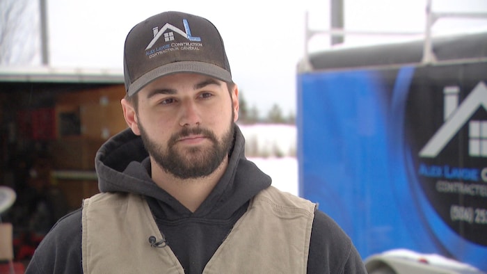 Alex Lavoie, coiffé d'une casquette au nom de son entreprise, est debout à l'extérieur, en hiver.