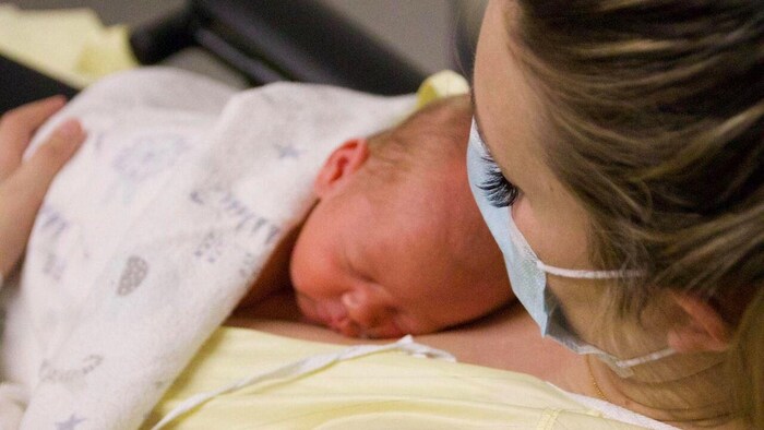 Alexa Forbes tenant son bébé contre elle. Elle porte un masque et une chemise d'hôpital.