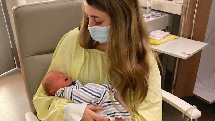 Masquée et vêtue d'une blouse d'hôpital, Alexa Forbes tient Keith dans ses bras, dans un fauteuil à l'hôpital.
