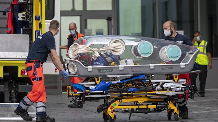 Deux ambulanciers poussent un caisson d'isolement placé sur une civière devant l'entrée d'un hôpital.
