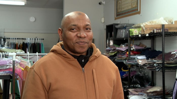 Un monsieur regarde vers la caméra et sourit. Il est debout dans un magasin devant des étagères et des étalages de vêtements bien remplis.