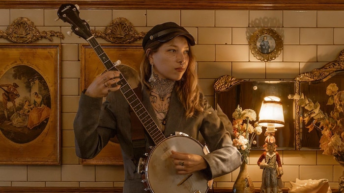 La femme joue du banjo devant un mur rempli de tableaux et de bibelots. 
