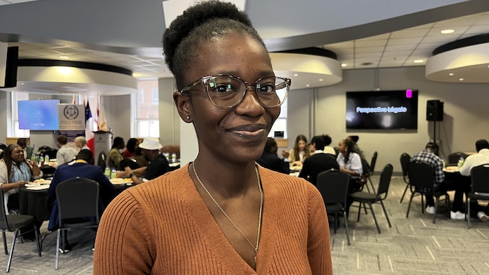 Une femme debout, de trois quarts, portant des lunettes et un chandail orange, sourit en posant pour une photo dans une salle de conférence avec des dizaines de personnes en arrière-plan.