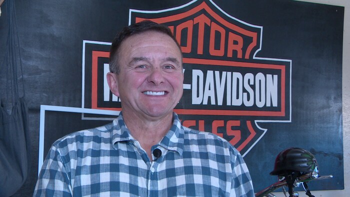 Un homme pose pour une photo et sourit à belles dents devant une affiche d'Harley-Davidson.