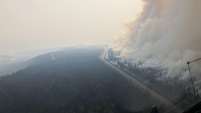 Depuis le poste de pilotage d'un hélicoptère, une route traverse des collines boisées noyées de fumée; un panache s'élève à droite de la chaussée.