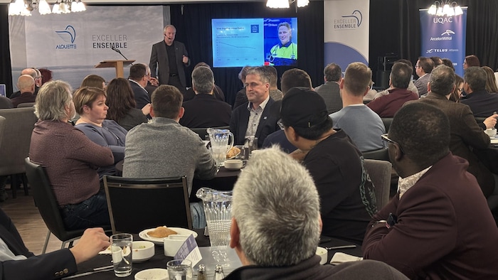 Des gens assis dans une salle de congrès écoutent un homme portant un micro.