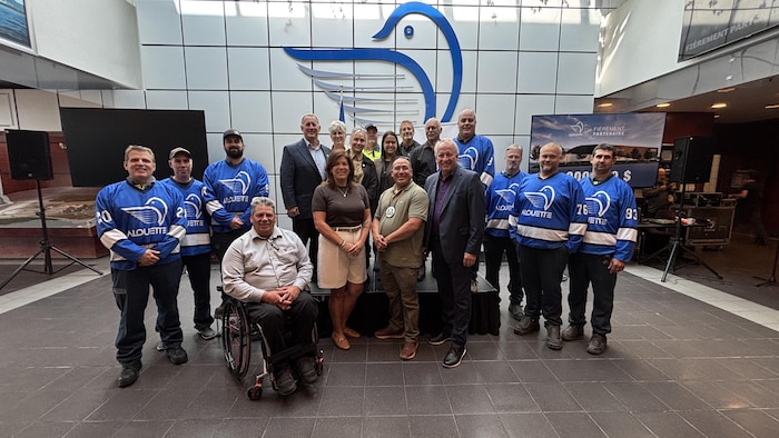 Photo de groupe large dans l'atrium moderne de l'aluminerie, sous un grand logo d'oiseau. Au centre, des dignitaires en tenue de ville sont entourés de plusieurs hommes portant des chandails de hockey Alouette.
