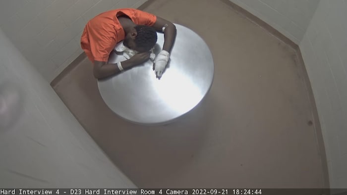 Un homme en tenue de prisonnier orange avachi sur une table dans une salle d'interrogatoire.