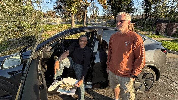 Un homme assis dans sa voiture avec les plans d'une maison et un autre debout avec des lunettes de soleil devant un terrain vacant. 