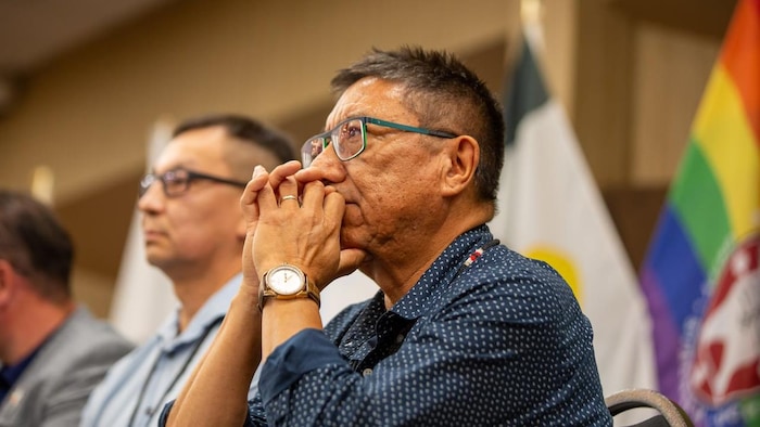 Un homme écoute attentivement un discours, les bras croisés, avec des drapeaux derrière lui.
