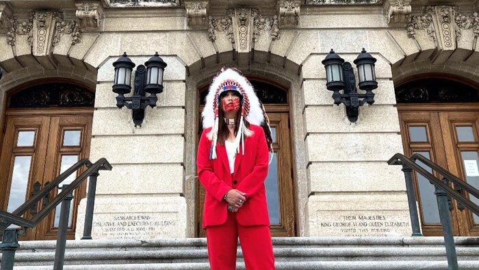 Aly Bear, vêtue d'une coiffe de chef et d'un complet veston rouge, debout devant l'escalier d'un édifice.