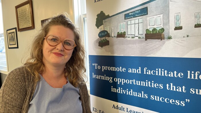 Une femme pose pour une photo le long d'un mur décoré d'une affiche annonçant un programme d'éducation aux adultes.