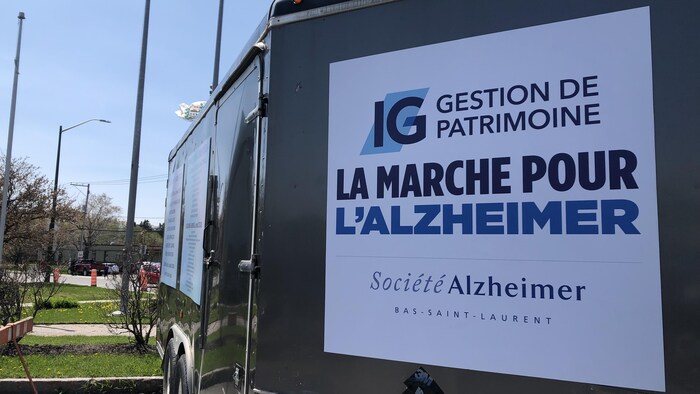 Sur une remorque fermée, une affiche avec l'inscription "La marche pour l'alzheimer", Société Alzheimer Bas-saint-Laurent. En arrière plan, le ciel, le drapeau du Québec et le drapeau du Canada.