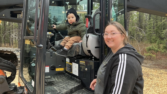 Un bambin assis dans une machine forestière, une main sur le manche de commande, regarde la caméra. Sa maman, debout juste à côté de la machine, sourit à la caméra.