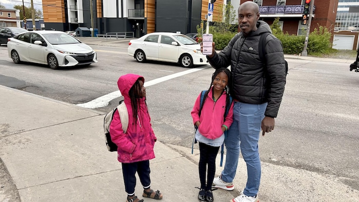 Sur le trottoir, un père accompagne ses feux fillettes. Il présente l'écran de son téléphone à la caméra.