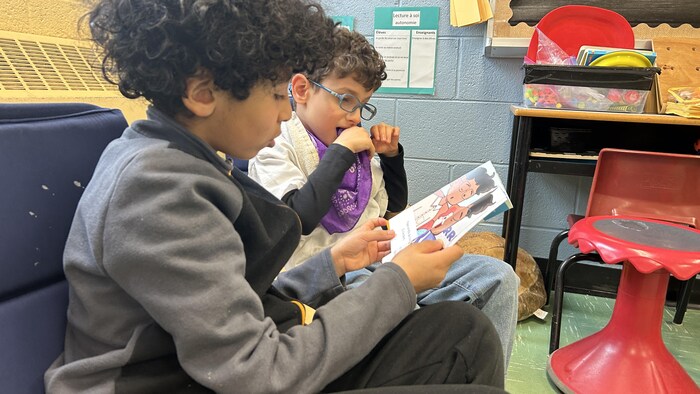 Abdou est assis près d'Amir sur des fauteuils dans la classe.  Il tient un livre dans ses mains.