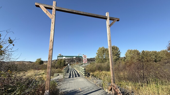 Une arche en bois à l'entrée d'un sentier. On voit un pont et une usine plus loin.