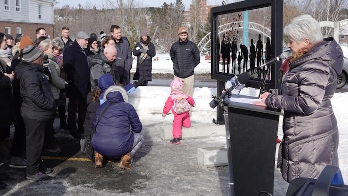 Sylvie Blanchette se tient debout derrière un micro, avec derrière elle la structure commémorative, qu'une foule de personnes contemple.