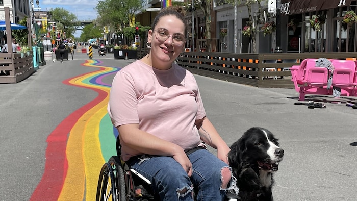 Anaïs Sabourin se déplace en fauteuil roulant en compagnie de sa chienne d'assistance, Mika. Ils sont sur la rue Ste-Catherine. Des arc-en-ciel ont été dessinés sur la rue.