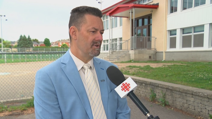 André Bernier speaks to a microphone in front of a school. organizations concerned about "unacceptable compressions"