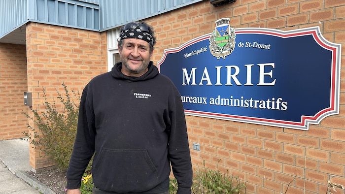 André Lechasseur pose devant l'affiche de la mairie de Saint-Donat.