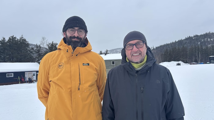 Deux hommes souriants posent côte à côte dans un paysage hivernal enneigé. L’un porte un manteau jaune et une tuque noire, l'autre un manteau noir. En arrière-plan, on aperçoit des bâtiments, une forêt de conifères et un ciel couvert.