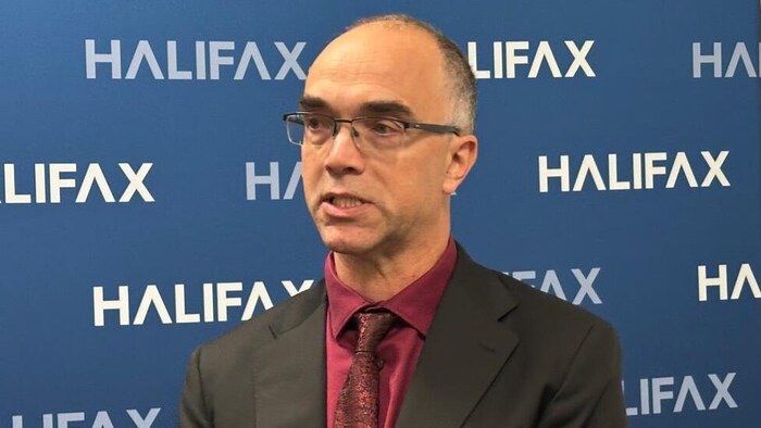 Un homme mince, portant des lunettes, vêtu d'un complet, devant un panneau sur lequel figure le logo de la Municipalité régionale d'Halifax.