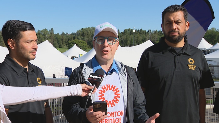 Andrew Knack avec une casquette et un t-shirt ayant le logo du Festival du patrimoine, avec deux hommes à ses côtés devant un micro.