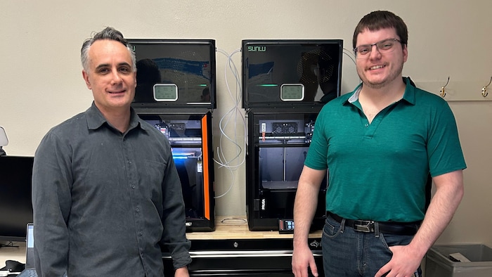 Deux hommes posent debout pour une photo, devant deux imprimantes 3D, et sourient.