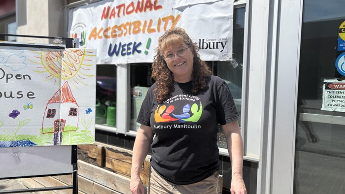 Angela Gray porte un chandail avec le nom de son organisme qui y est inscrit et pose devant une affiche qui mentionne que c’est la semaine nationale de l’accessibilité.