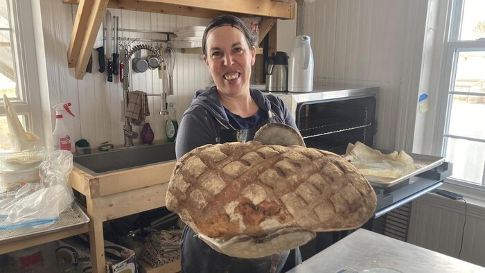 Une femme tient une énorme miche dans ses mains. Un four se trouve derrière elle, dans une cuisine de boulangerie. 