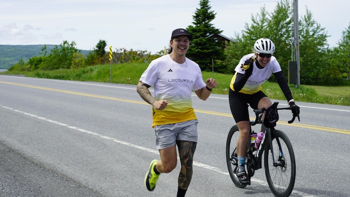 Un coureur de l'équipe et une cycliste qui les accompagne sourit à la caméra. Le coureur fait du mieux qu'il peut pour sourire malgré la fatigue.