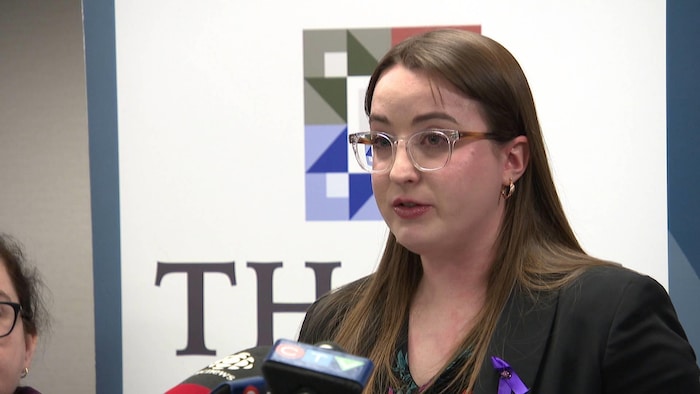 Une femme de profil, debout devant des micros, parle en conférence de presse. Elle porte un ruban violet.