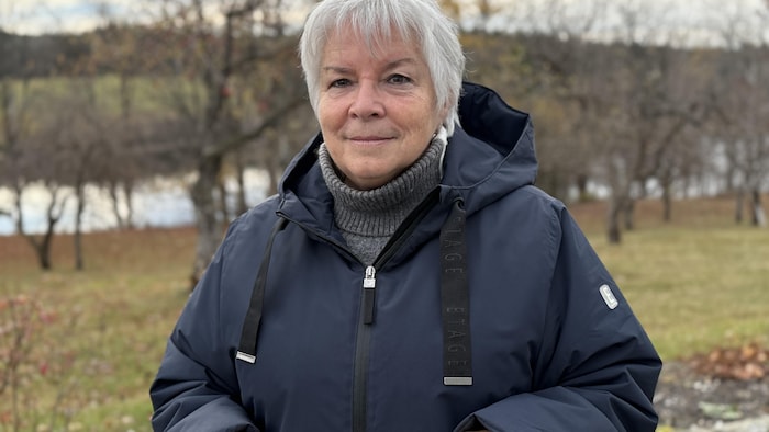 Annie Godbout, vêtue d'un imperméable, devant un plan d'eau, regarde la caméra,
