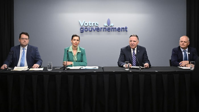 Quatre ministres devant un mur gris sur lequel on peut lire « Votre gouvernement ». 